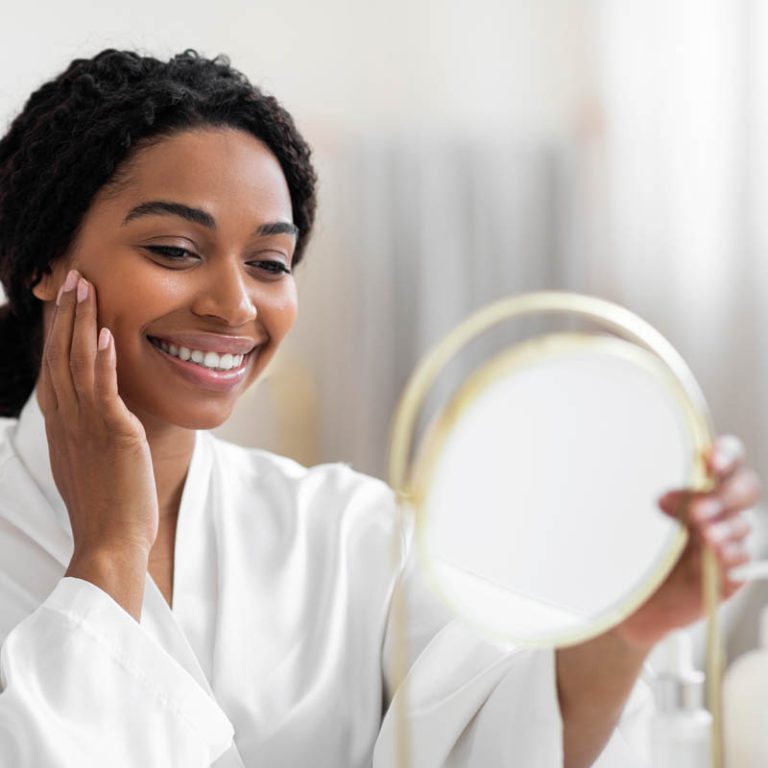 Attractive Young Black Woman Looking At Mirror And Touching Perfect Skin On Face While Making Beauty Care Treatments At Home, Pretty African American Lady Smiling To Her Reflection, Closeup