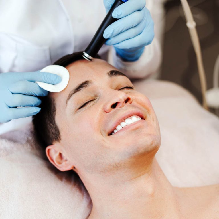 Profession cosmetologist making vacuum facial skin cleaning for smiling man in beauty salon. Handsome male lying with closed eyes. Cosmetology procedure concept